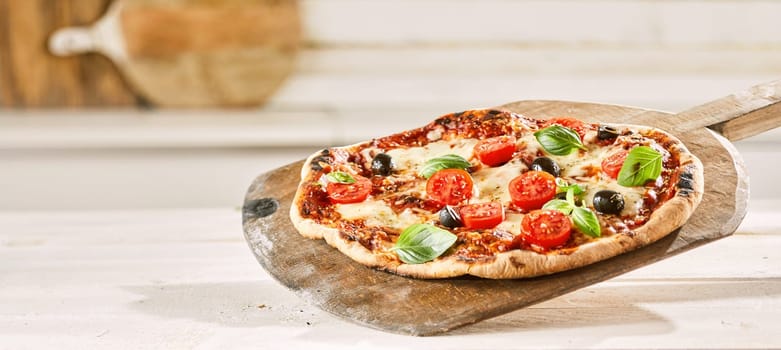 Margherita Italian pizza with extra trimmings topped with fresh tomato, olives and shaved parmesan cheese served on an old wooden pizzeria paddle in panorama banner format