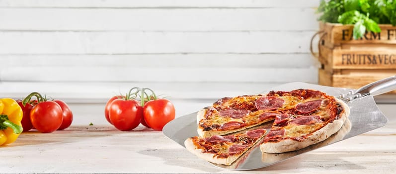 Panorama banner with a traditional Italian salami pizza served on a metal paddle with a slice removed and fresh ingredients and herbs on a rustic white wood background