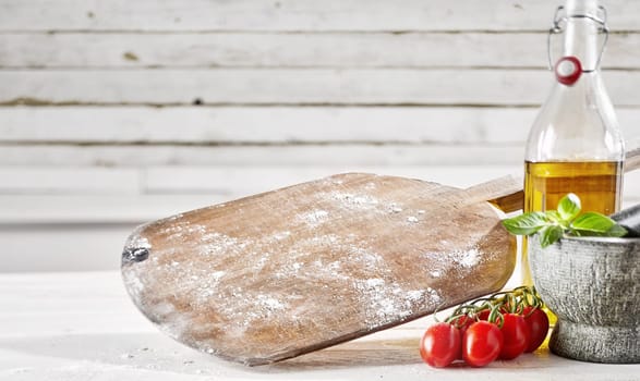 Old wooden empty pizzeria pizza paddle with flour on a white wood table alongside tomatoes, olive oil and a pestle and mortar with copy space