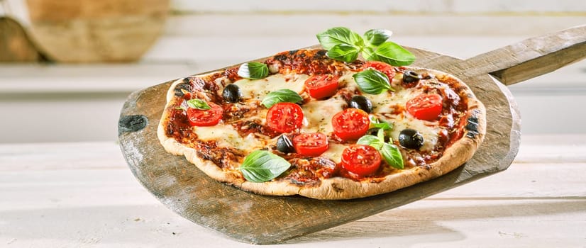 Margherita pizza with tomato, olives and fresh basil leaves on a thin crispy flame-grilled base served on an old wooden paddle at a pizzeria