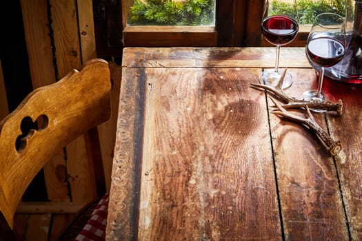 Old bare rustic wooden chair and table with deer antlers and two glasses of red wine in front of the window for food or product placement