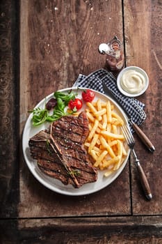 Grilled lean T-bone steak with golden potato chips and salad served with a dip or dressing on a rustic wooden table in a top down view with copy space