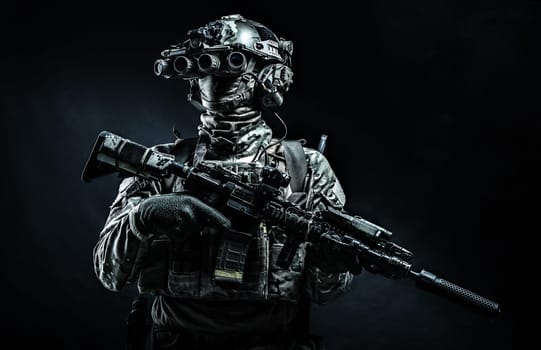 Army infantry in battle uniform, armed assault rifle with laser sight and silencer, standing in darkness, looking through night vision goggles, low key half length, studio portrait on black background