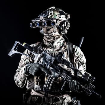 Marine rider in camouflage uniform and face mask, patrolling in darkness with quad-tube night vision goggles on battle helmet, holding modern assault rifle, low key studio portrait on black background
