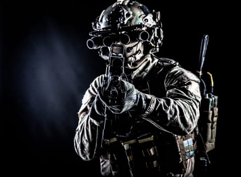 Soldier of special forces in camo combat uniform, load carrier, helmet, equipped night sight goggles, tactical radio headset, aiming assault rifle with collimator sight in camera, low key studio shoot