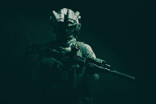 Half length, low key studio shoot of army soldier, marine infantryman in mask, camo uniform, equipped modern ammunition, armed service rifle standing in darkness with night vision device on helmet