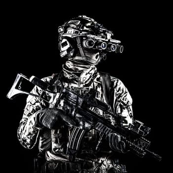 Marine rider in camouflage uniform and face mask, patrolling in darkness with quad-tube night vision goggles on battle helmet, holding modern assault rifle, low key studio portrait on black background