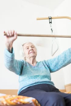 Elderly 96 years old woman exercising with a stick sitting on her bad. Geriatric health care home assisted support for older people concept. Care for the elderly.