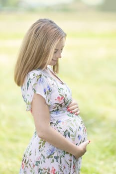 Portrait of beautiful pregnant woman in white summer dress relaxing in meadow full of yellow blooming flovers. Concept of healthy maternity care.