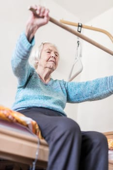 Elderly 96 years old woman exercising with a stick sitting on her bad. Geriatric health care home assisted support for older people concept. Care for the elderly.
