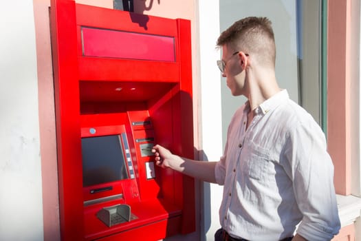 young man student withdrawing money from a bank cash point, outdoors