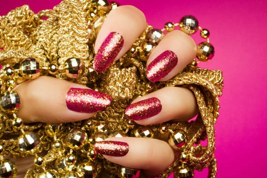 Female hand with gold glittered pink nails is holding gold pearl jewelry on pink background, nail care and manicure concept.