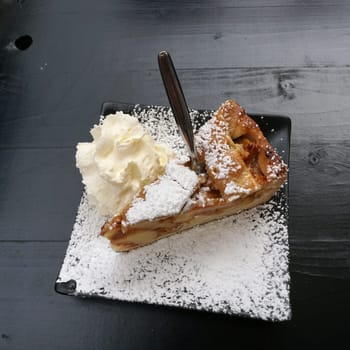 a piece of apple pie with cream and powdered sugar on a black, quatrefoil plate
