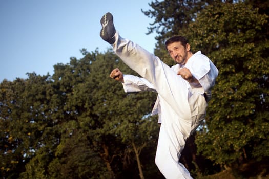 The karateist in white kimono is kicking high and practicing martial arts outdoors in nature.