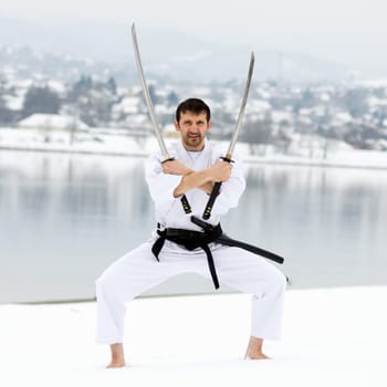 Man in white kimono and black belt is practicing martial arts with two samurai swords barefoot on snow at winter.