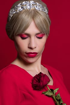 The beautiful woman in red dress with tiara is holding red rose on red background.