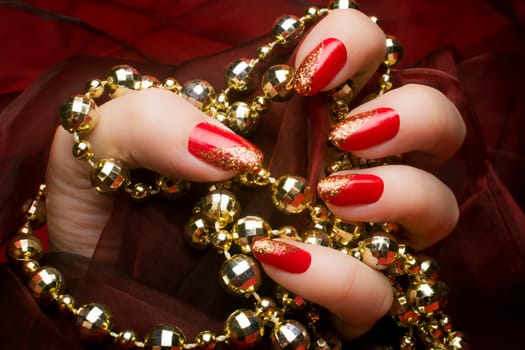 Female hand with golden glittered red nails is holding a gold pearl jewelry on dark red background.