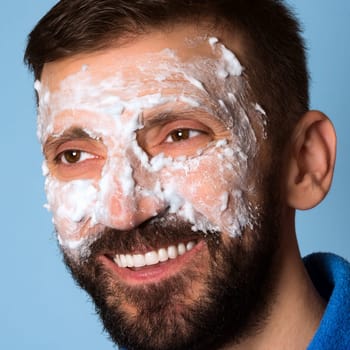 The smiling middle aged man is using foam for skincare on his face on blue background.