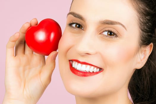 The smiling beautiful woman with red lips is holding little red heart in hand on pink background.