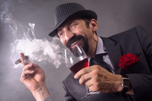 The smiling winking elegant gentleman in suit and hat is smoking cigar and drinking wine on gray background.