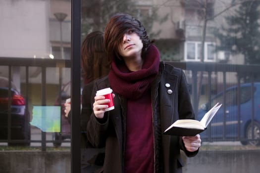 The urban style student boy with book and coffee is standing outdoors at the street and resting.