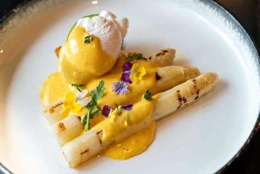 Gourmet grilled white asparagus with poached egg serve with hollandaise savory sauce decorate with edible colorful flowers