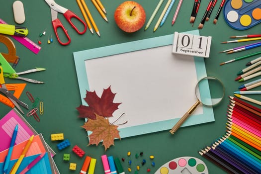Blue frame with blank sheet, autumn leaves, stationery accessories on green background. Top view, copy space. School accessories for children's education and development. Day of knowledges
