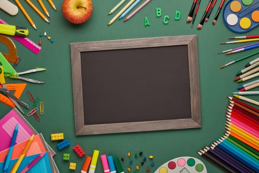 Blank blackboard, apple and stationery accessories on green background. Top view, copy space. School accessories for children's education and development. Office supplies. Preparation for school