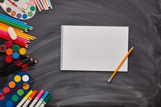 Blank sheet of writing-book and stationery accessories on background of school blackboard. Top view, copy space. School accessories for children's education and development. Art lesson or drawing