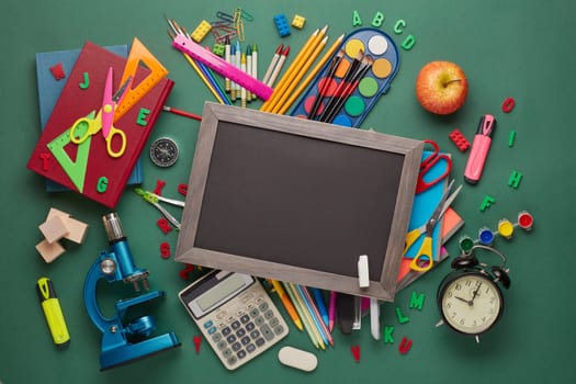 Blank blackboard, microscope and stationery accessories: pencils, pens, other office supplies on green background. Top view, copy space. School accessories for children's education and development