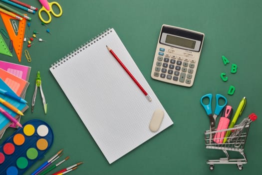 Blank sheet of writing-book, shopping trolley and stationery accessories for children's education on green background. Top view, copy space. Preparation for school, planning budget. Cost of education