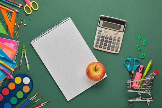 Blank sheet of writing-book, shopping trolley and stationery accessories for children's education on green background. Top view, copy space. Preparation for school, planning budget. Cost of education