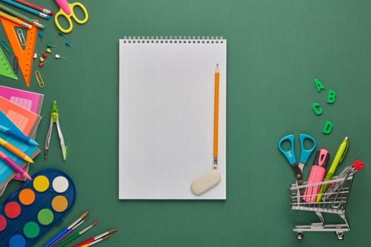 Blank sheet of writing-book, shopping trolley and stationery accessories on green background. Top view, copy space. School accessories for children's education. Office supplies. Preparation for school