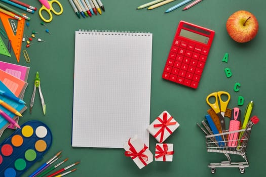Blank writing-book, shopping trolley, boxes with purchases, stationery accessories for education on green background. Top view, copy space. Preparation for school, planning budget. Cost of education