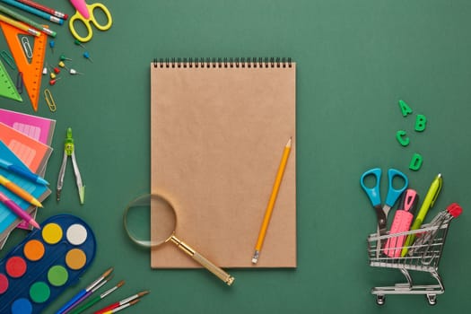 Blank sheet of writing-book, shopping trolley and stationery accessories on green background. Top view, copy space. School accessories for children's education. Office supplies. Preparation for school