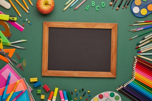 Blank blackboard, apple and stationery accessories on green background. Top view, copy space. School accessories for children's education and development. Office supplies. Preparation for school