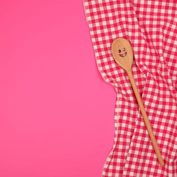 wooden spoon with a carved face on a red kitchen towel, pink background, copy space