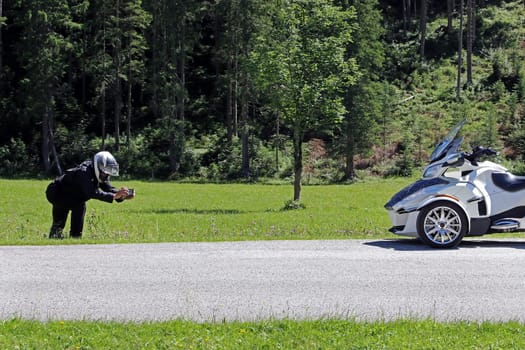 A man wearing motorcycle clothing and a safety helmet photographs his Spyder with a smartphone.