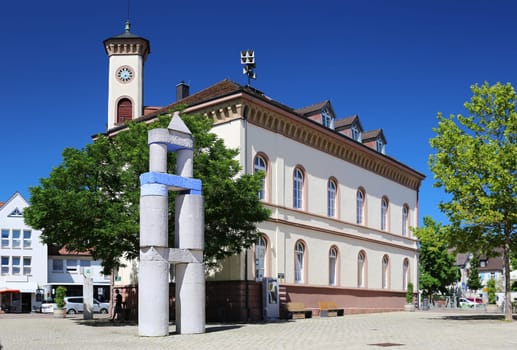 Pedestrian area in the city center of Müllheim, called Markgräfler Platz. In the foreground you can see the sculpture "The city gate"", behind it is the building of the Land Registry with tower clock."