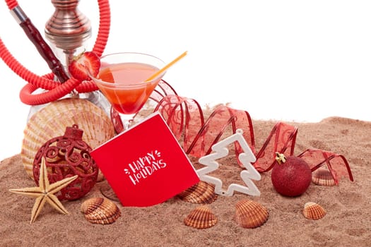 Festive beach still life: glass with cocktail, hookah (shisha), seashells, sand and christmas ornaments on white background. New Year and Christmas. Winter vacation in warm countries, beach holiday