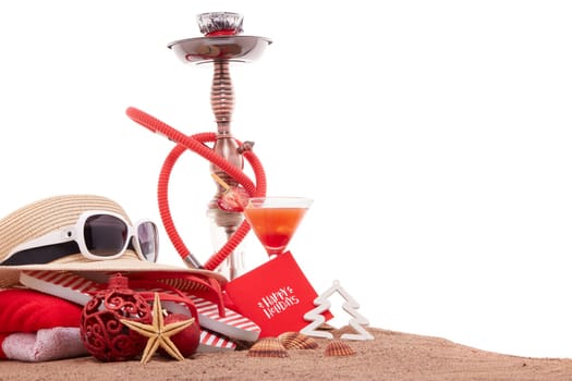 Beach accessories, seashells, sand, glass with cocktail, hookah (shisha) and christmas ornaments on white background. New Year and Christmas. Winter vacation in warm countries, beach holiday