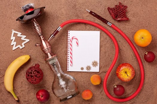 Hookah (shisha), fruit, blank sheet of notebook and christmas ornaments on sand background. Top view. New Year and Christmas. Winter vacation in warm countries, beach holiday