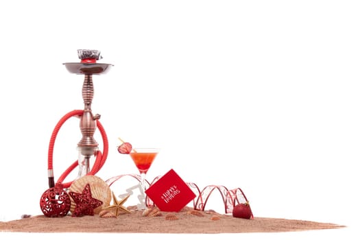 Festive beach still life: glass with cocktail, hookah (shisha), seashells, sand and christmas ornaments on white background. New Year and Christmas. Winter vacation in warm countries, beach holiday