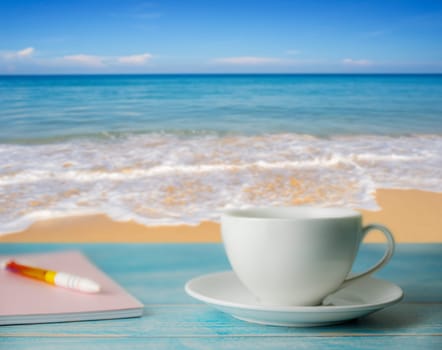 A cup of coffee and notebook with blurred beach sand and blue sky in day light, Relax and remember concept