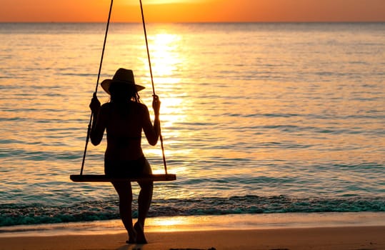 Silhouette woman wear bikini and straw hat swing the swings at the beach on summer vacation at sunset. Enjoying and relaxing girl on holiday. Summer vibes. Woman watching beautiful sunset sky.