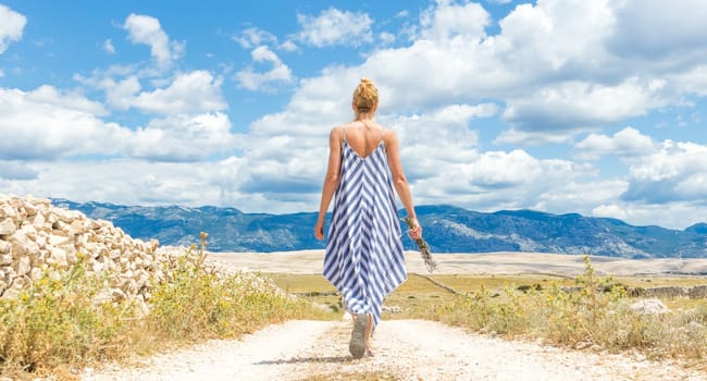 Rear view of woman in summer dress holding bouquet of lavender flowers while walking outdoor through dry rocky Mediterranean coast lanscape on Pag island, Croatia in summertime.