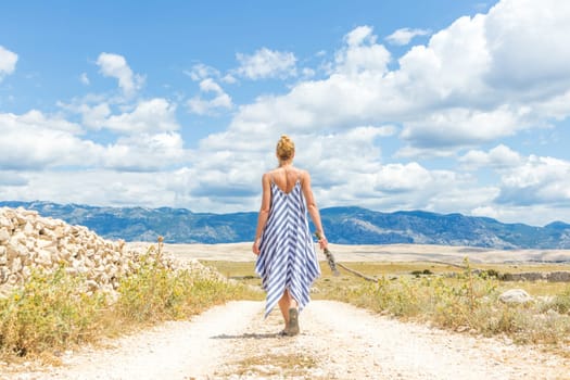 Rear view of woman in summer dress holding bouquet of lavender flowers while walking outdoor through dry rocky Mediterranean coast lanscape on Pag island, Croatia in summertime.