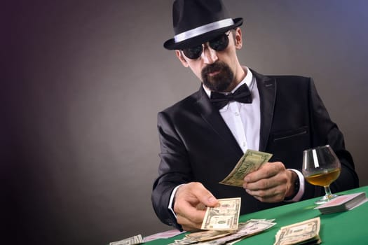 Elegant gentleman in tuxedo is gambling at the table on gray background.