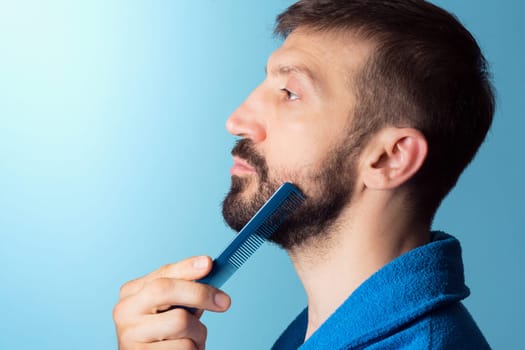 The middle aged man is combing his beard on blue background, man`s care concept.