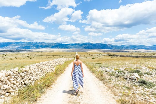 Caucasian young woman in summer dress holding bouquet of lavender flowers while walking outdoor through dry rocky Mediterranean Croatian coast lanscape on Pag island in summertime.